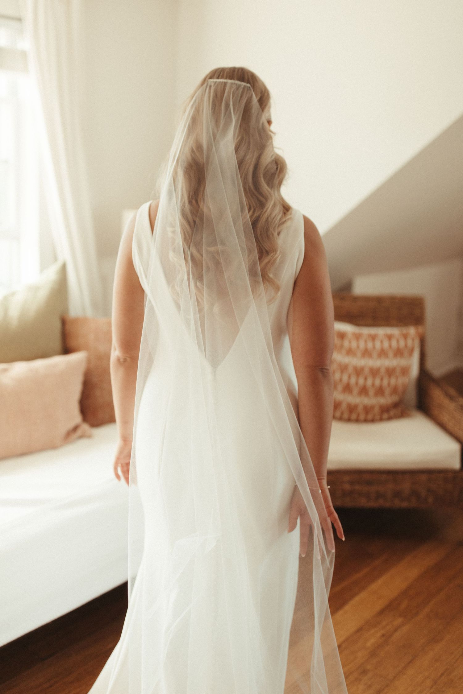 Back view of a bride in a flowing white wedding dress and veil standing in a sunlit room with decor.