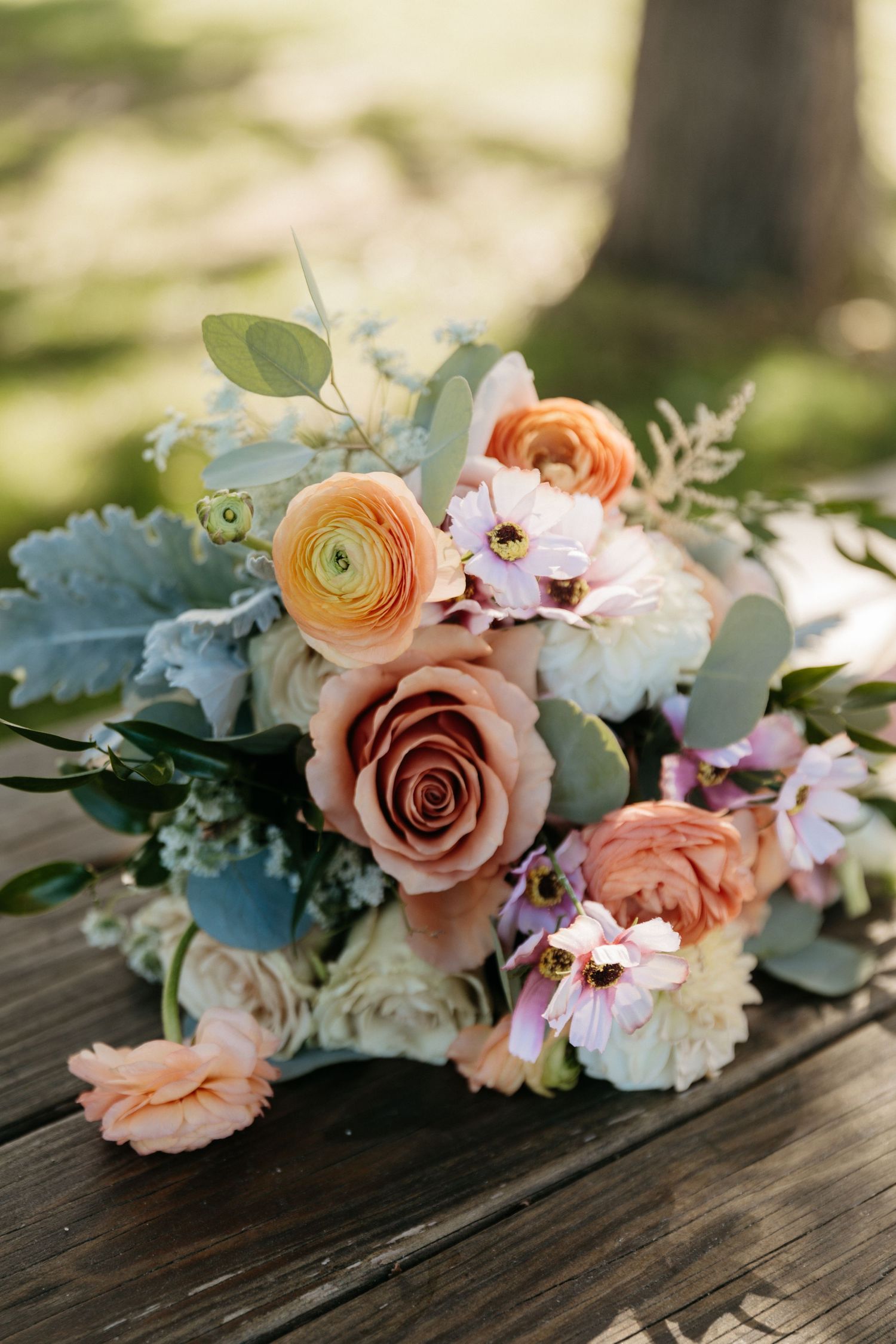 A romantic wedding bouquet with peach roses, ranunculus, and eucalyptus leaves on rustic wooden surface.