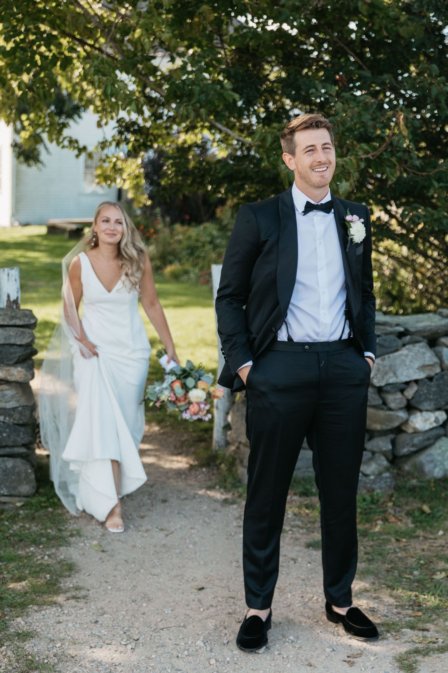Stylish wedding couple posing on garden path with stone wall backdrop.