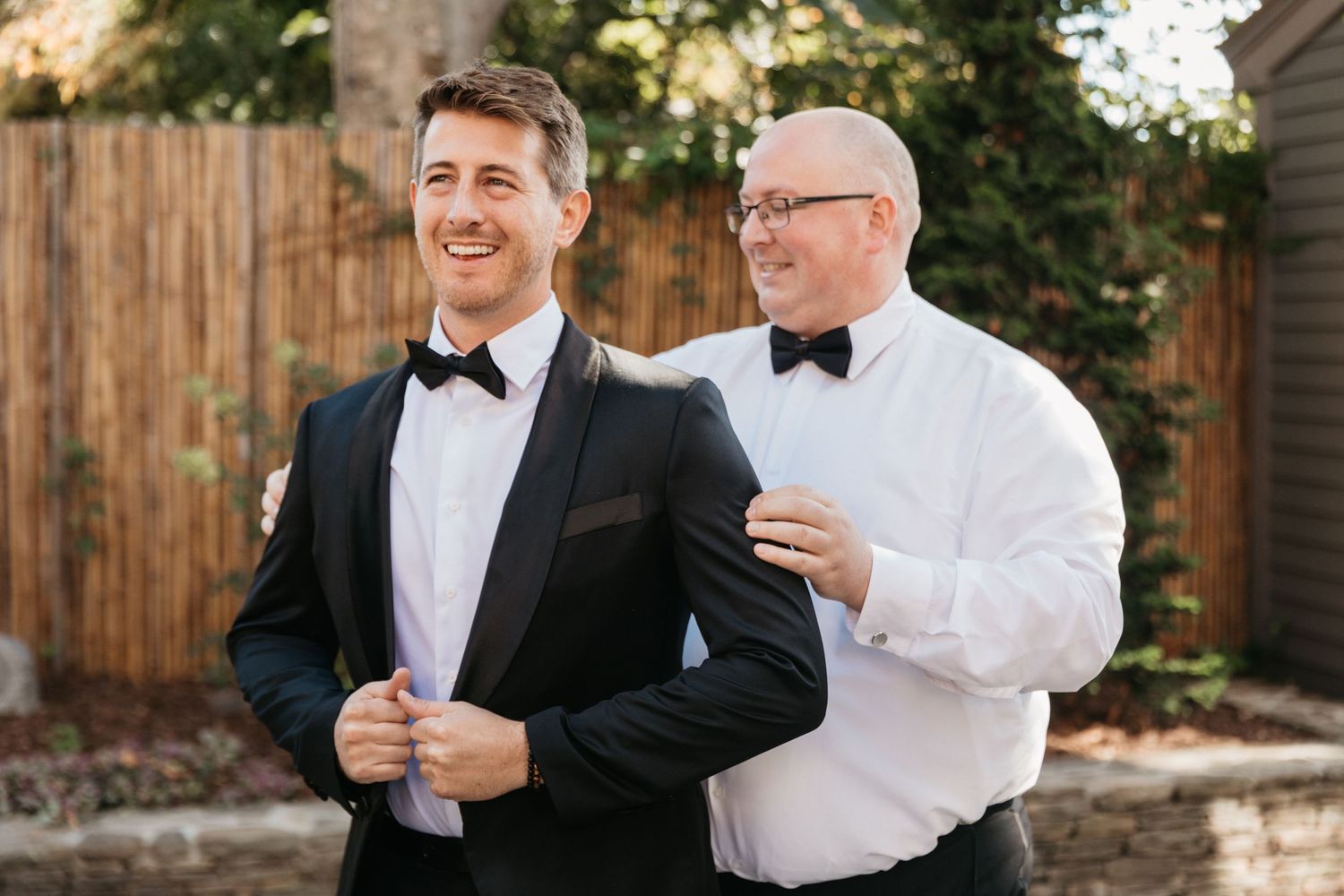Wedding party members in formal attire sharing a candid moment in front of wooden fence.