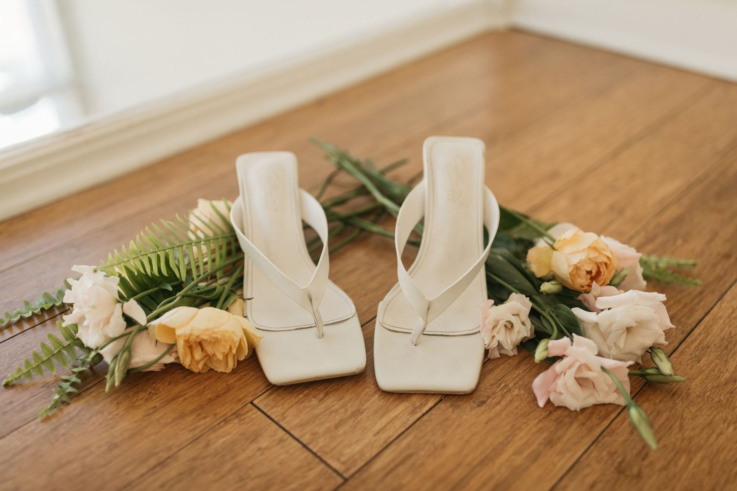 White square-toe bridal heels displayed with pink roses and greenery on wooden floor.