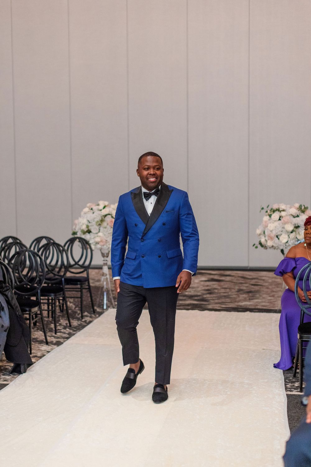 A groom walks down the aisle in a bright blue tuxedo jacket and black bow tie.