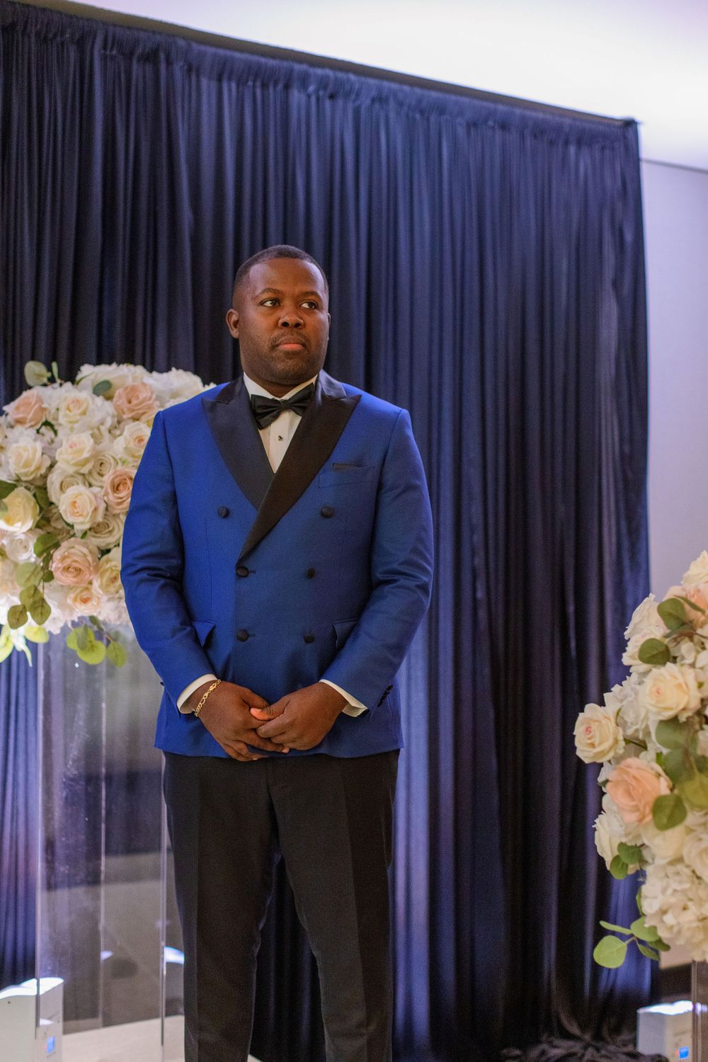A groom in a royal blue double-breasted tuxedo stands between floral arrangements.