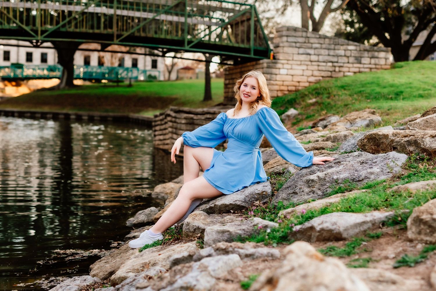 A woman in a blue dress poses along a riverbank with a bridge in the background.