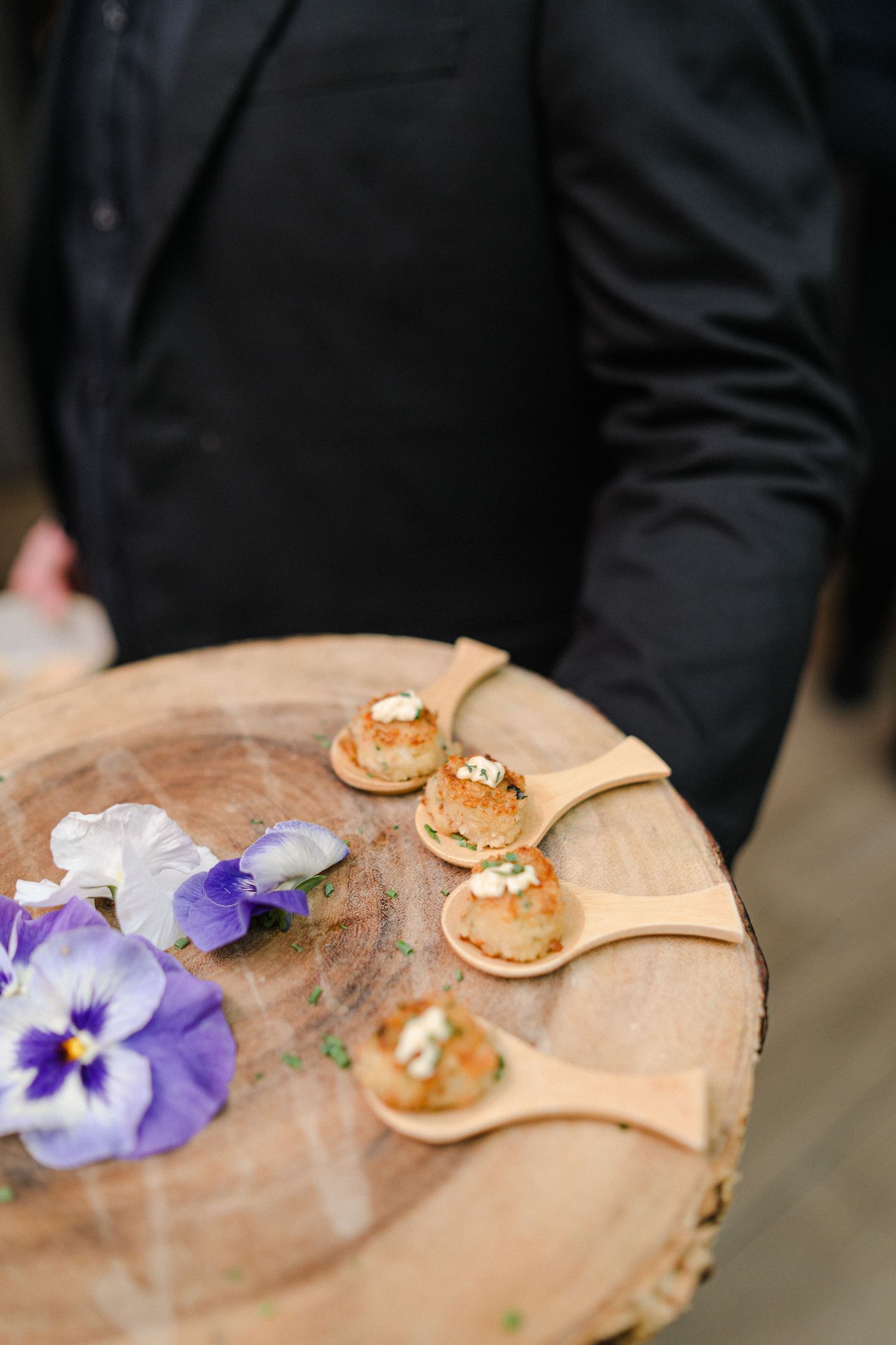 Wooden serving board with delicate appetizer spoons and purple pansies.