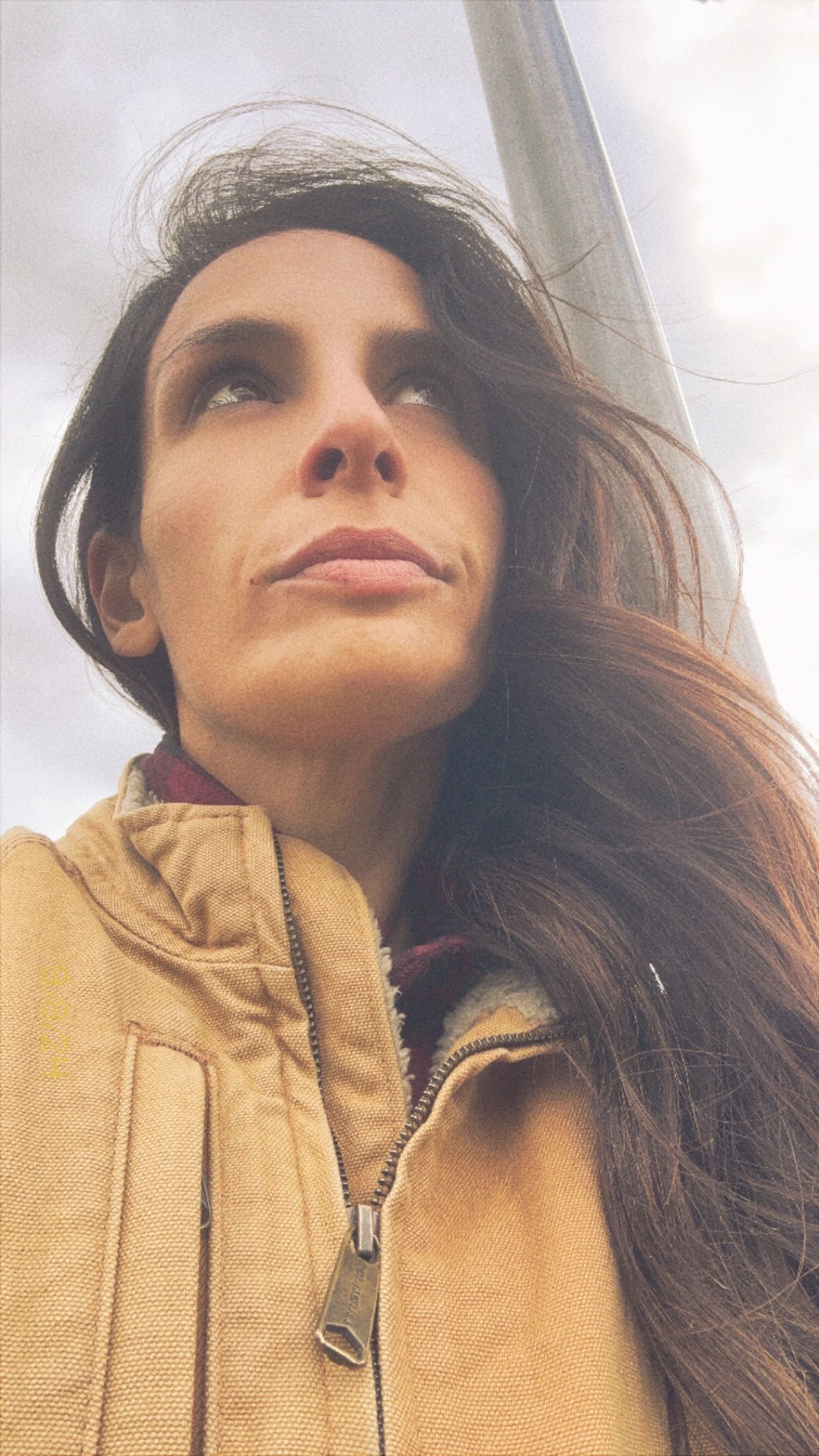 A person wearing a yellow hoodie gazes upward with windswept hair against a cloudy sky.