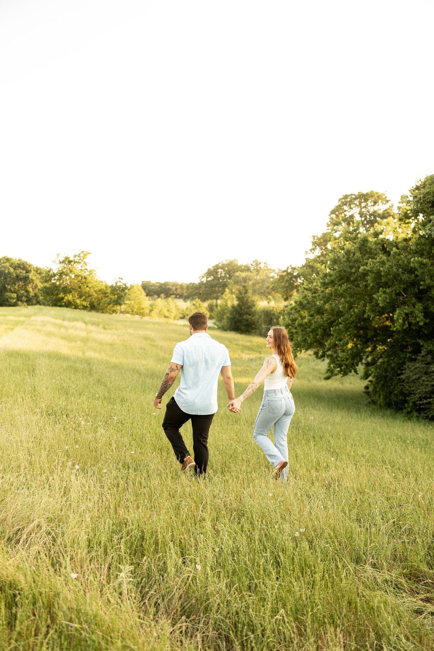 Two people hold hands while walking through a sunny meadow with tall grass and trees in the background.