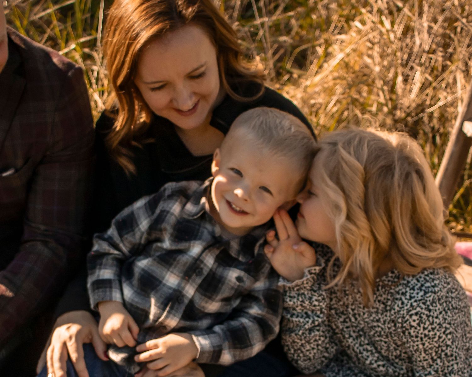 A mother holds her young son while his sister whispers a secret in his ear. All are smiling.