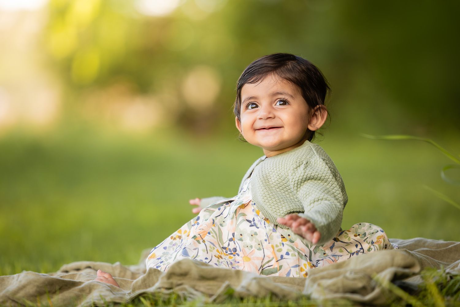 Baby in light green outfit sitting on hay bale in sunny garden setting.