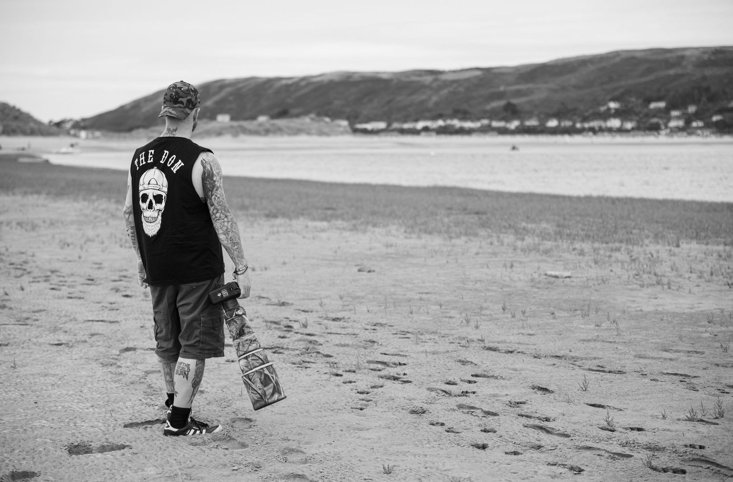 Tattooed man in skull tank top walking on sandy beach carrying skateboard in black and white.