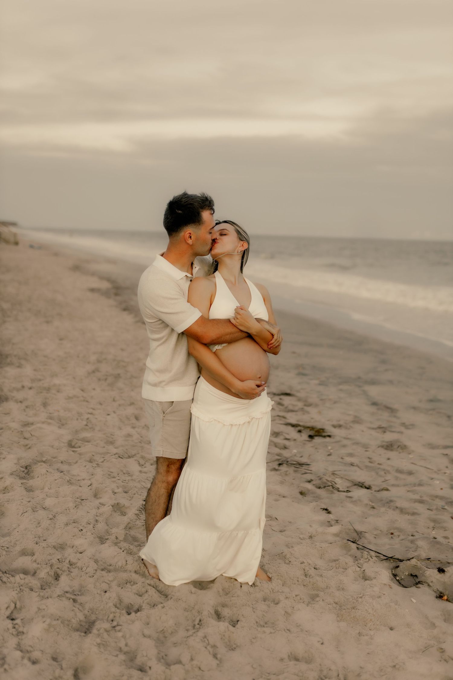 Romantic couple in white clothing embracing on a sandy beach at sunset.