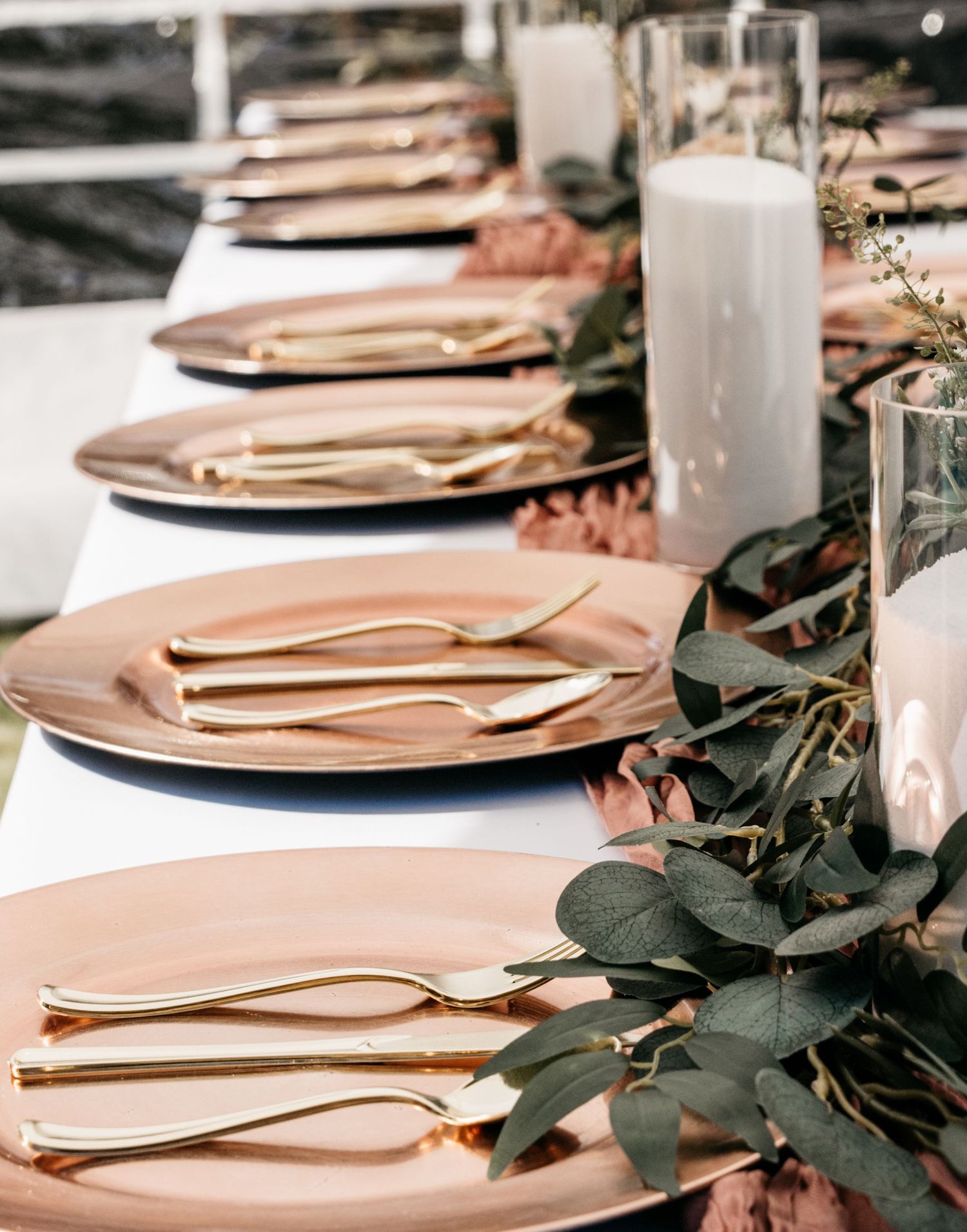 An elegant table setting with rose gold plates, gold flatware, and eucalyptus garland alongside tall white candles.