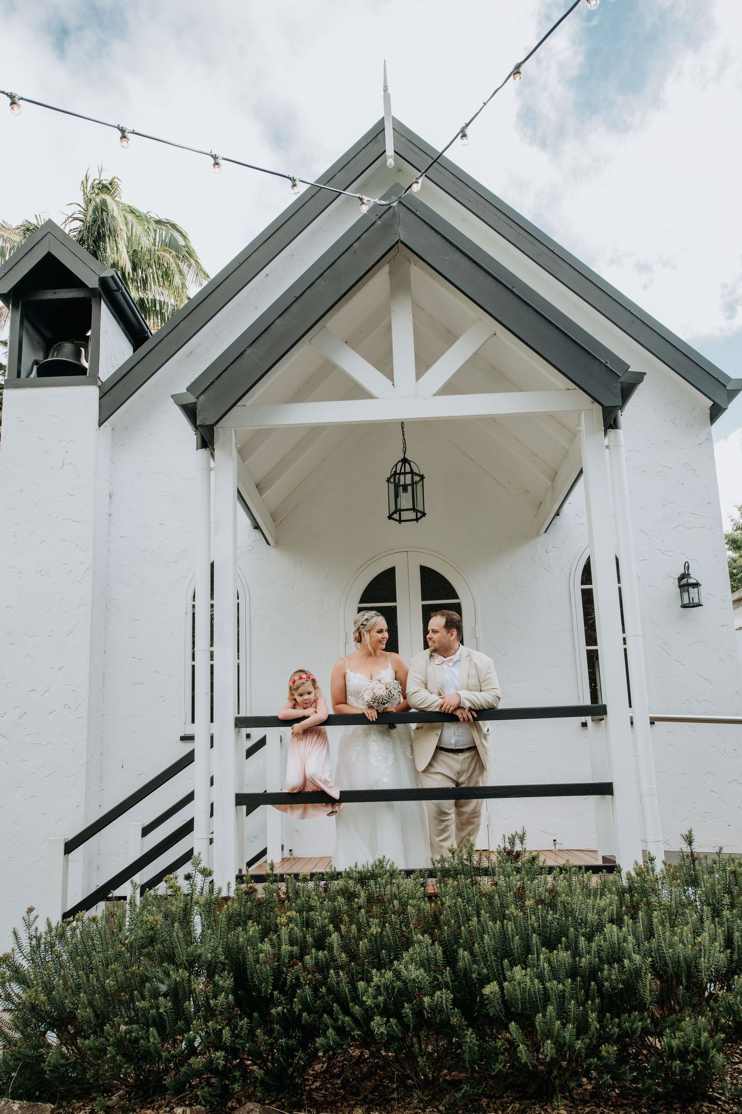 A charming white chapel entrance with a couple and flower girl standing on its porch during a wedding ceremony.