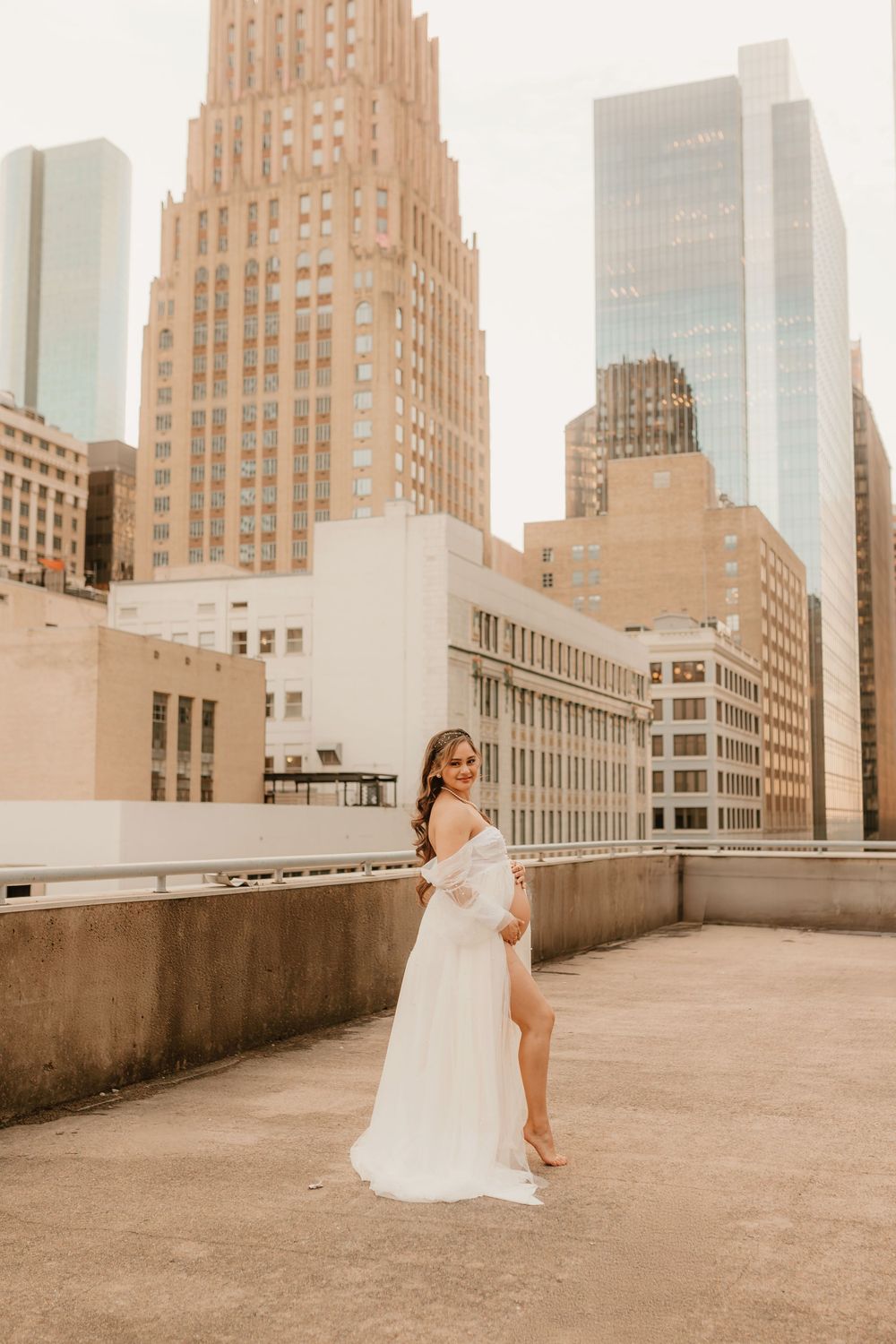 A solo maternity portrait in a white flowing dress on an urban rooftop during golden hour.