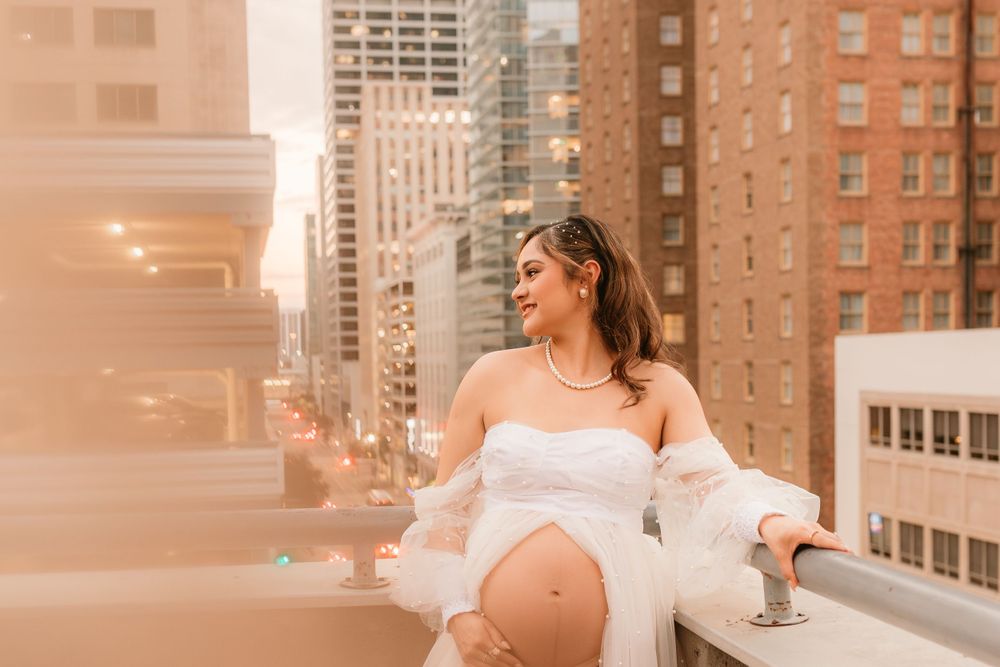 Expectant mother in white dress poses on rooftop with city skyline backdrop.