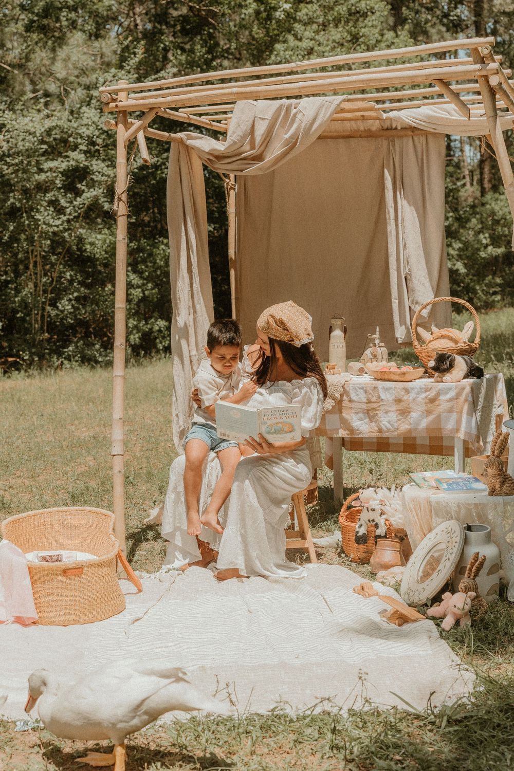 A cozy outdoor picnic setup with a bamboo canopy, white blanket, and wicker baskets in a sunny garden setting.