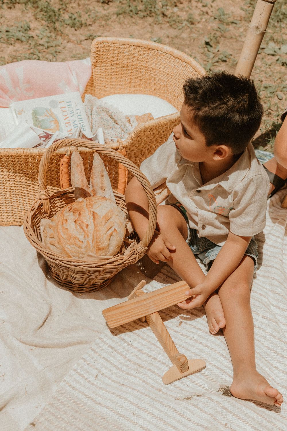 A child sits on a white blanket during a picnic with wooden toys and woven baskets in warm sunlight.