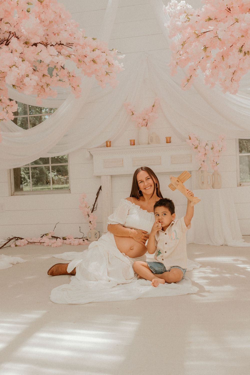 Dreamy maternity photo in white room with pink cherry blossoms and ethereal draping showing a mother and child on floor.