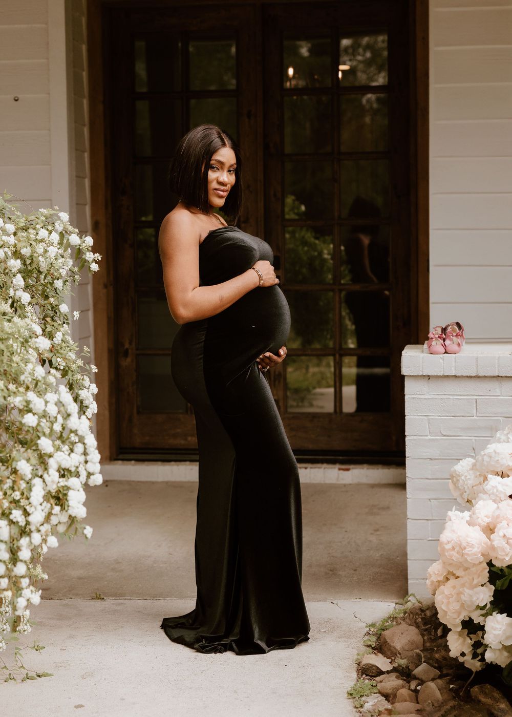 Elegant maternity photo in black gown with blooming flowers and doorway backdrop.