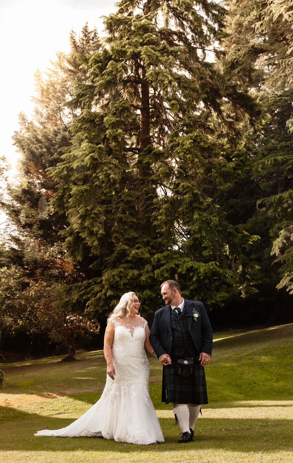 A couple walks together on a scenic grassy hill surrounded by tall evergreen trees during a golden sunset wedding photo session.