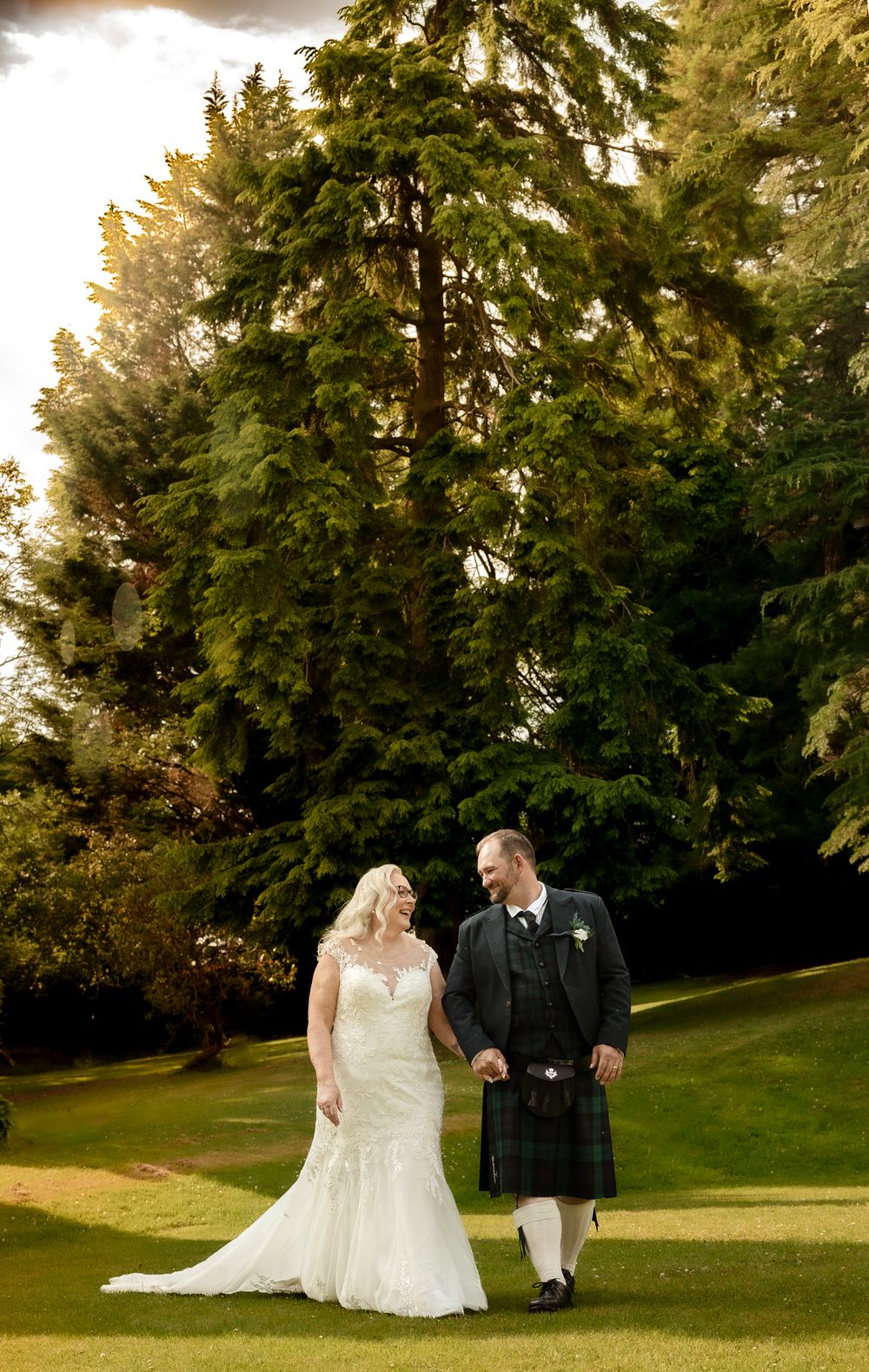 A couple walks hand in hand on a lush green lawn surrounded by tall evergreen trees at golden hour.