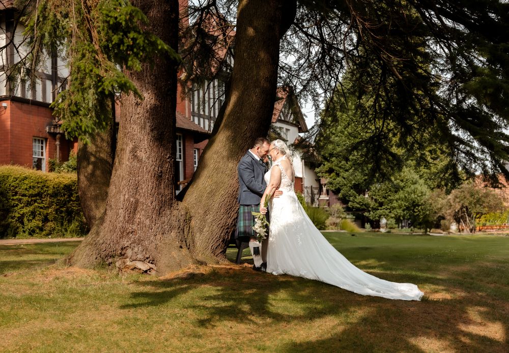 A couple in wedding attire stands together under a large tree on a manicured lawn near a brick building.