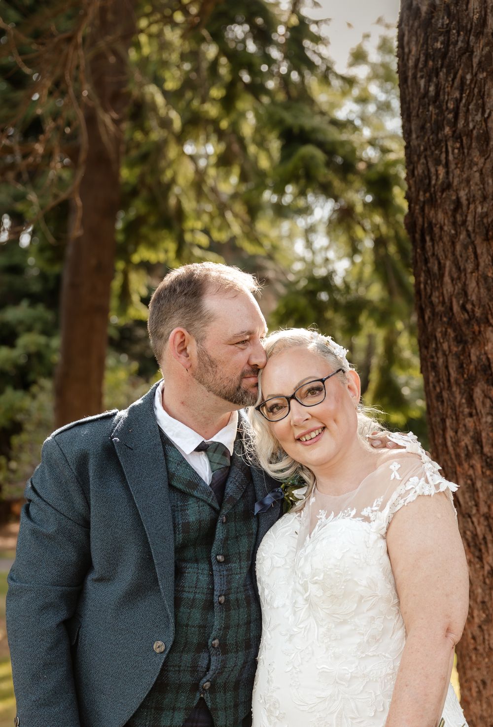 A happy couple embraces outdoors in a wooded setting, wearing formal wedding attire including a white dress and tartan vest.
