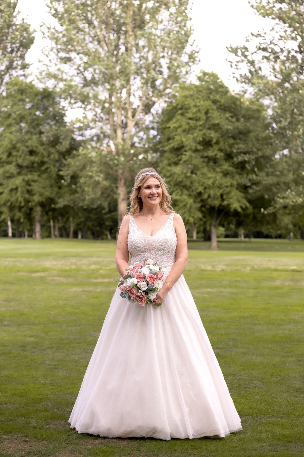 Woman in flowing white wedding gown stands in lush garden setting holding pink and white floral bouquet.