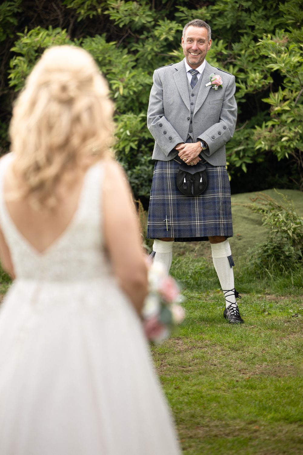 A traditional Scottish groom wearing a grey plaid kilt and formal jacket poses outdoors among green foliage.
