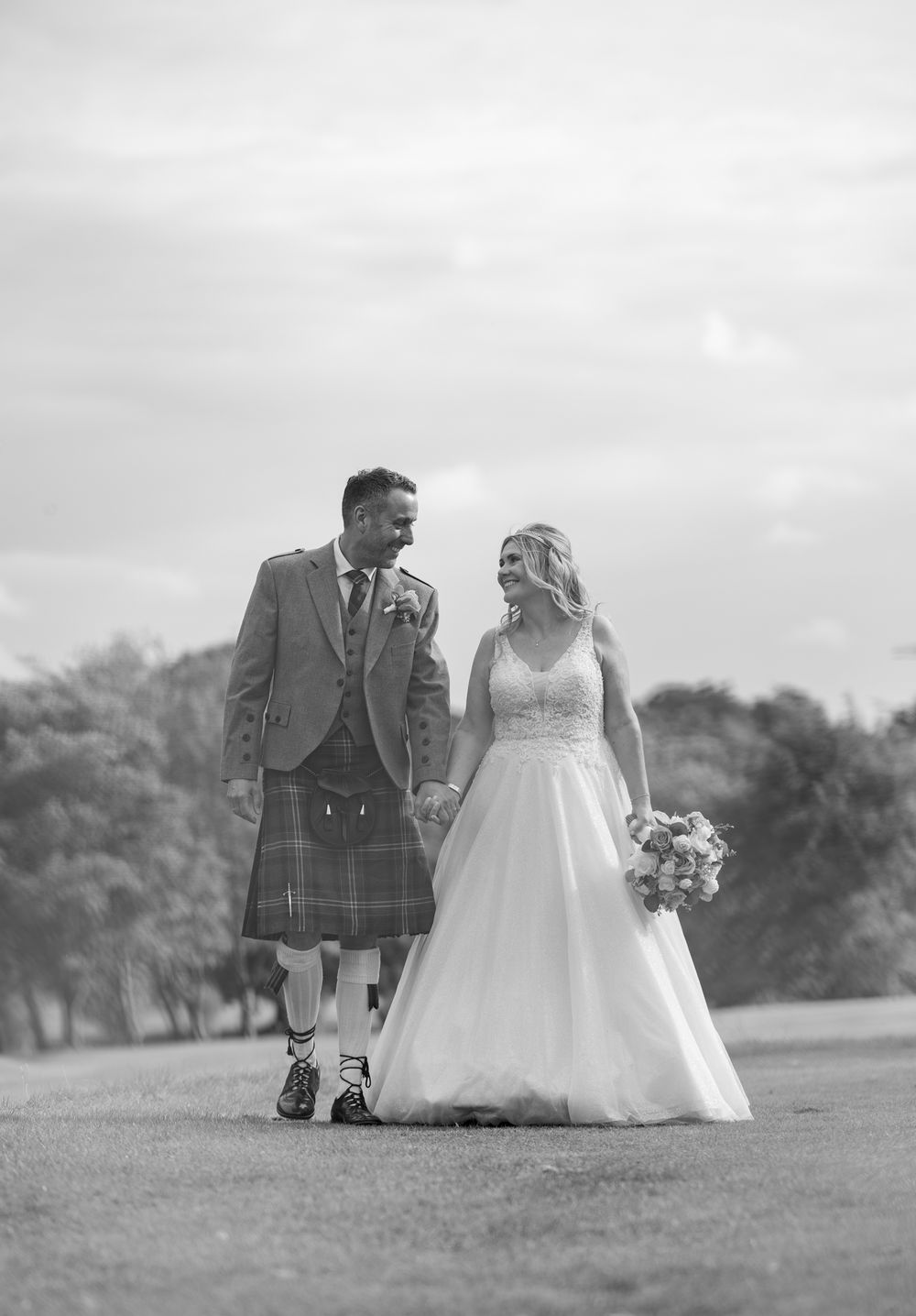 A black and white photograph of a couple at their Scottish wedding, with the groom wearing a traditional kilt on a cloudy day.