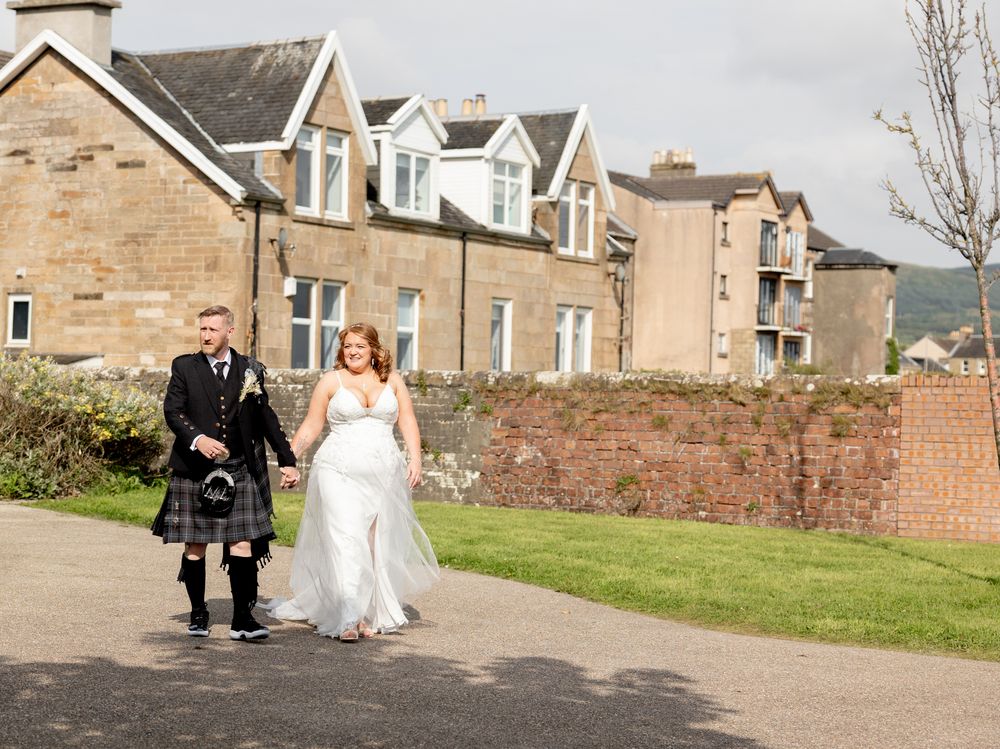 A couple walks together on a path outside traditional Scottish stone buildings, with the groom wearing a kilt for their wedding.