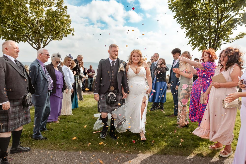 Wedding guests celebrate outside throwing confetti as people gather for an outdoor celebration on a sunny day.