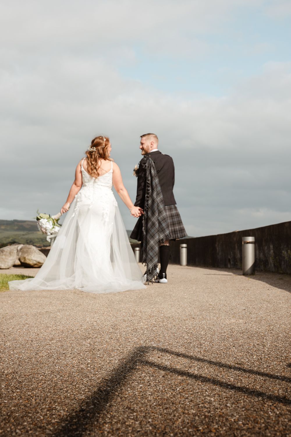 A bride in a white gown and groom in traditional Scottish kilt walk along a path under cloudy skies.