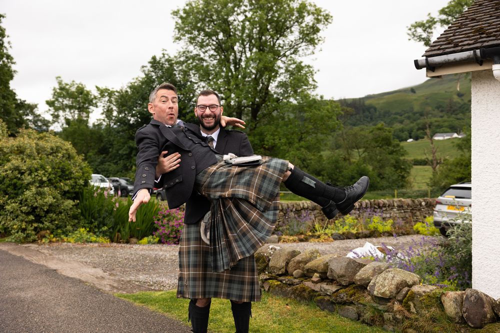 Two people in kilts sharing a joyful moment outside in a scenic Scottish countryside setting.