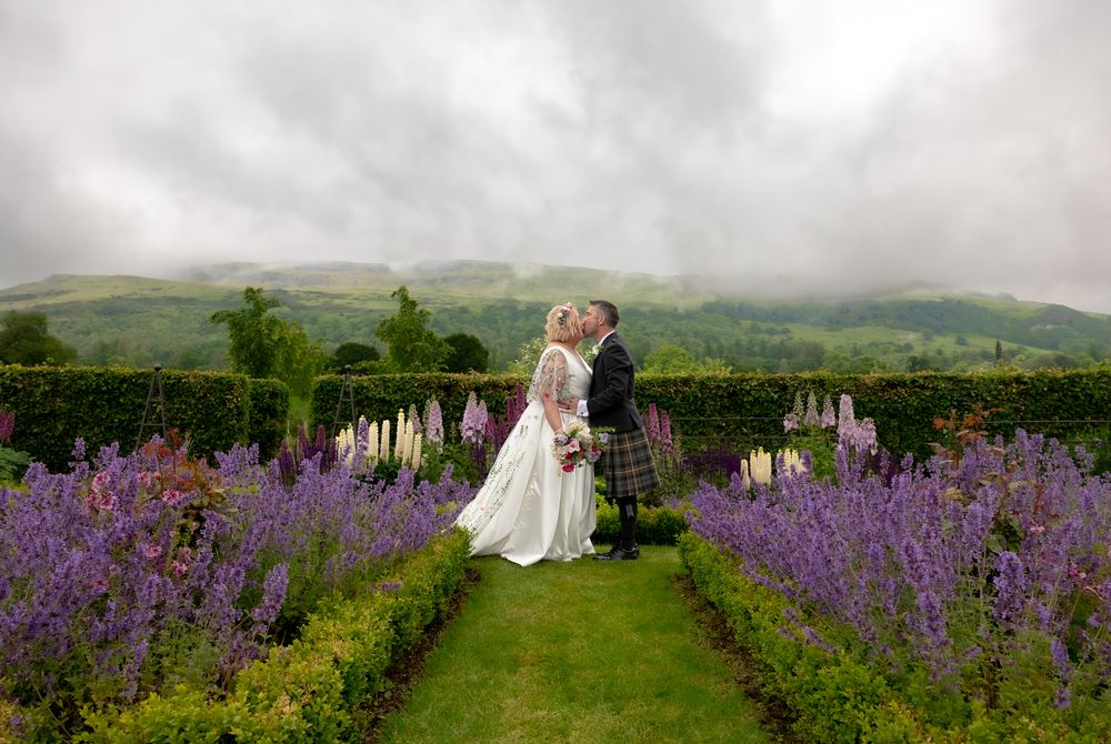 A couple embraces in a scenic garden path lined with vibrant purple lupines against rolling Scottish hills in the misty background.