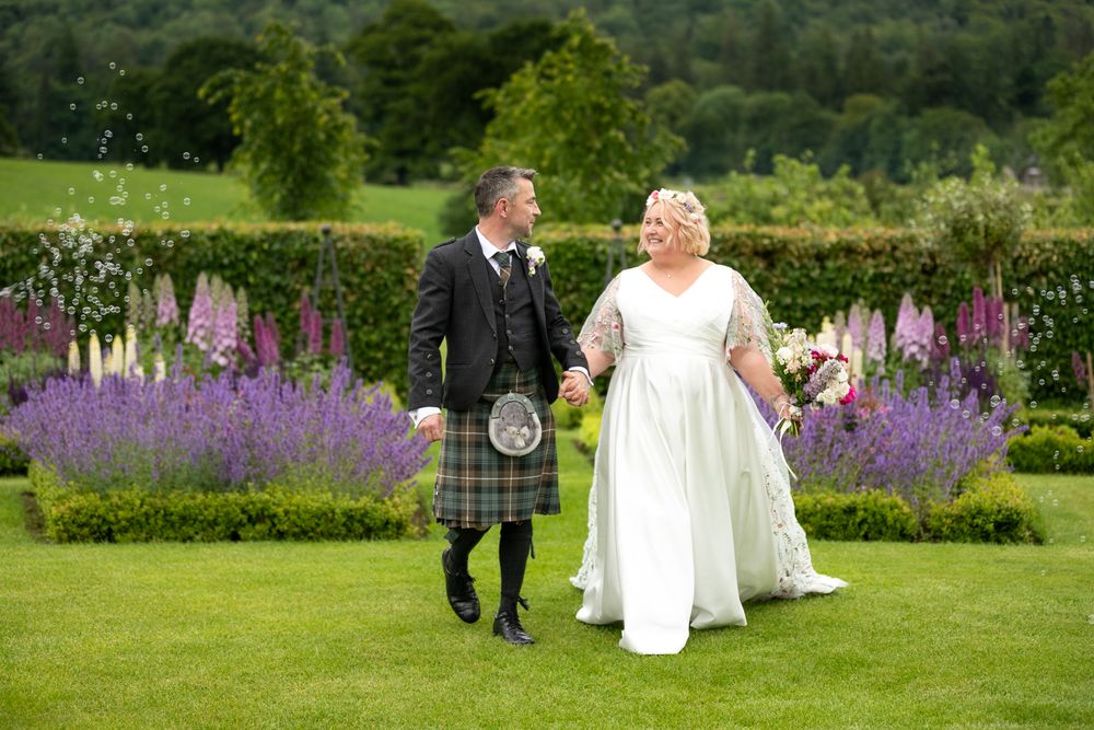 A wedding couple walks together across a lush green lawn with vibrant purple flowers blooming in the garden behind them.