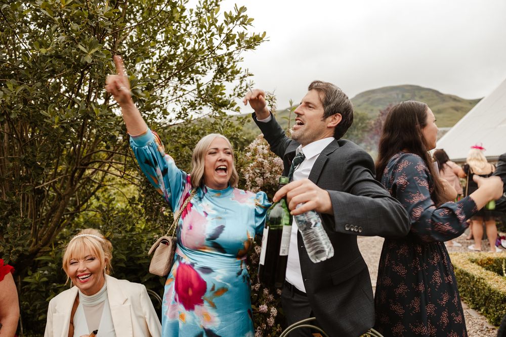 Wedding guests enthusiastically celebrate outside with raised drinks against a backdrop of scenic mountains at an outdoor reception.