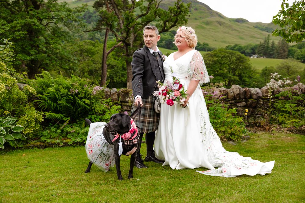 Wedding couple poses outdoors in green garden setting with two dogs wearing matching accessories at Scottish venue.