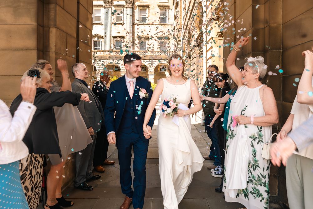 Wedding guests shower the newlyweds with confetti as they exit a grand stone building together.