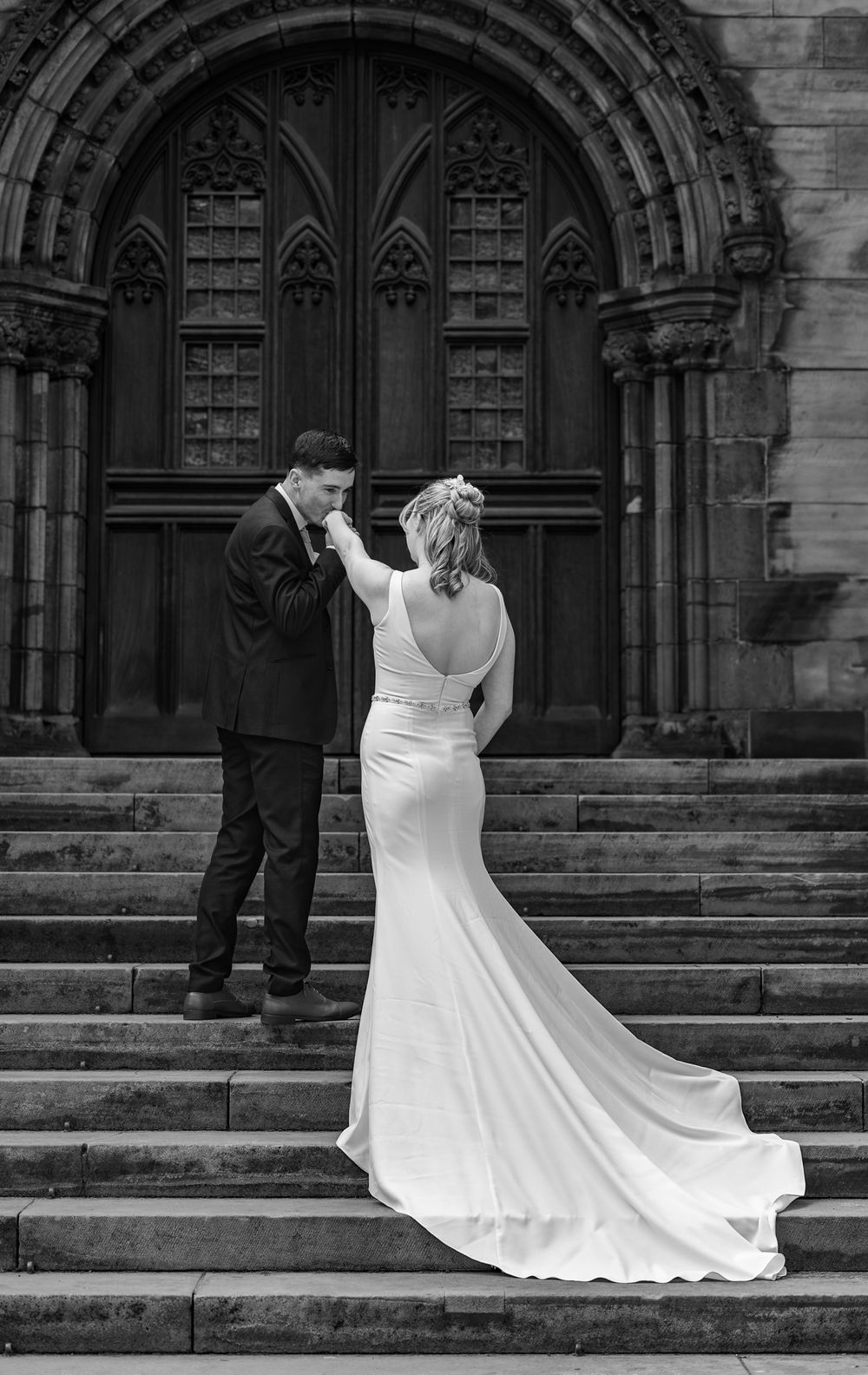A bride in a flowing white dress ascends stone church steps while her dress train gracefully cascades behind her.