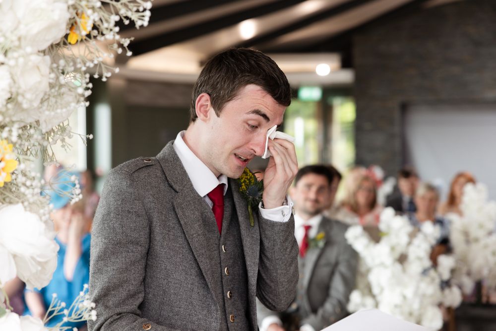 An emotional groom in a brown suit wipes his eye during a wedding ceremony surrounded by white floral decorations.