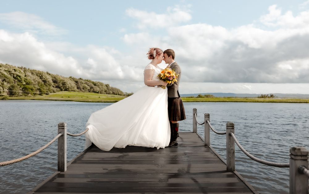 A couple in wedding attire embrace on a wooden dock overlooking a serene lake and hills under cloudy skies.