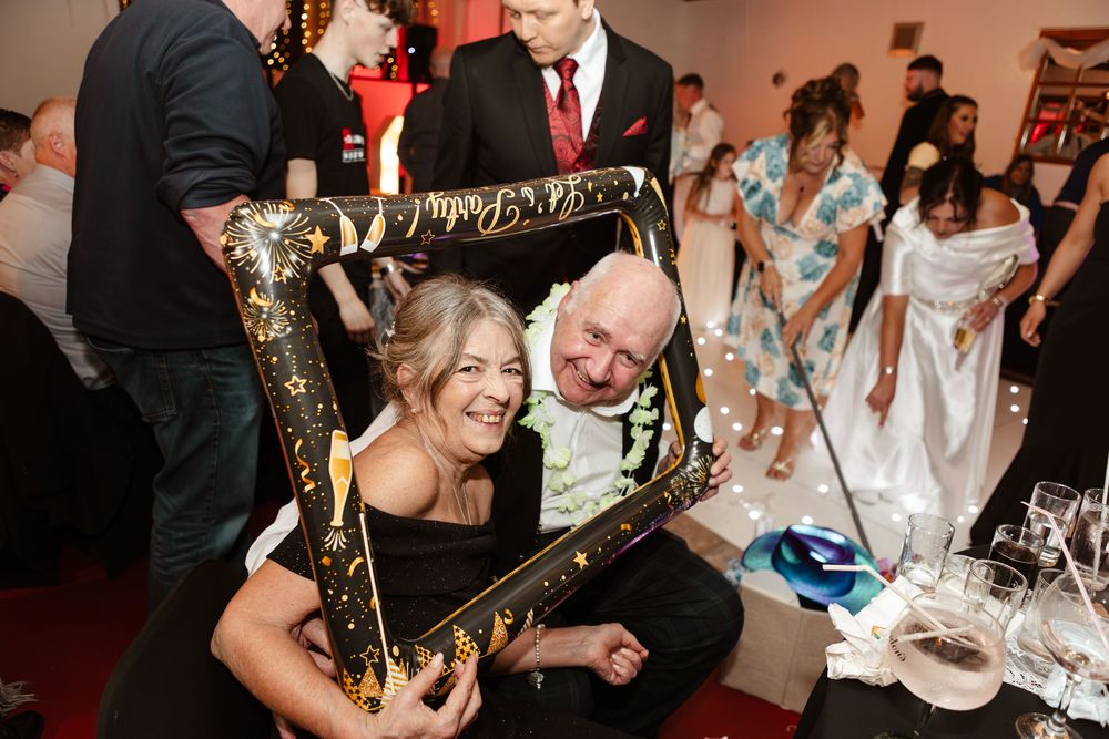 Two people share a joyful moment while posing with an ornate gold frame at a wedding reception.