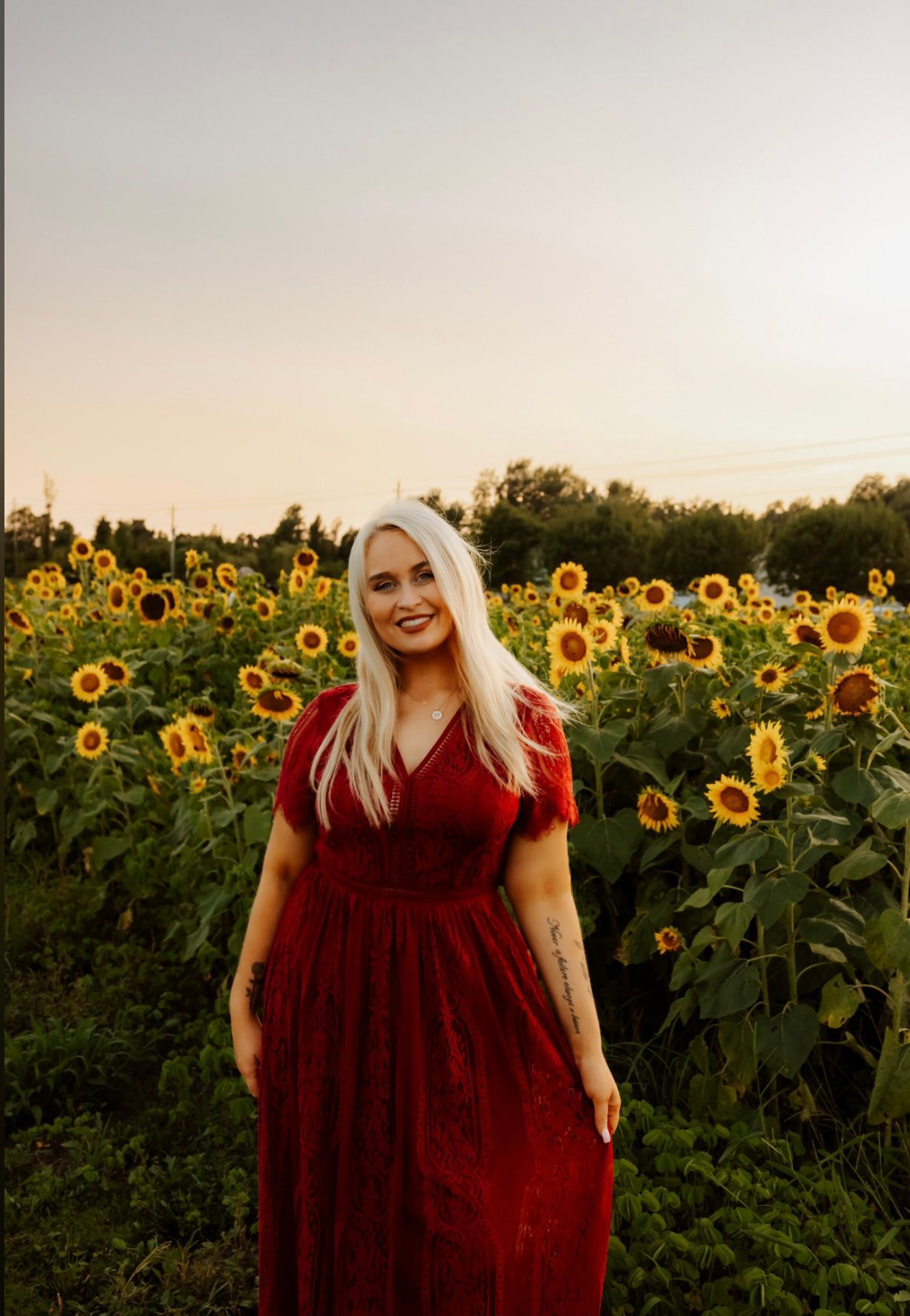 Woman in red dress smiling while standing in blooming sunflower field at sunset.