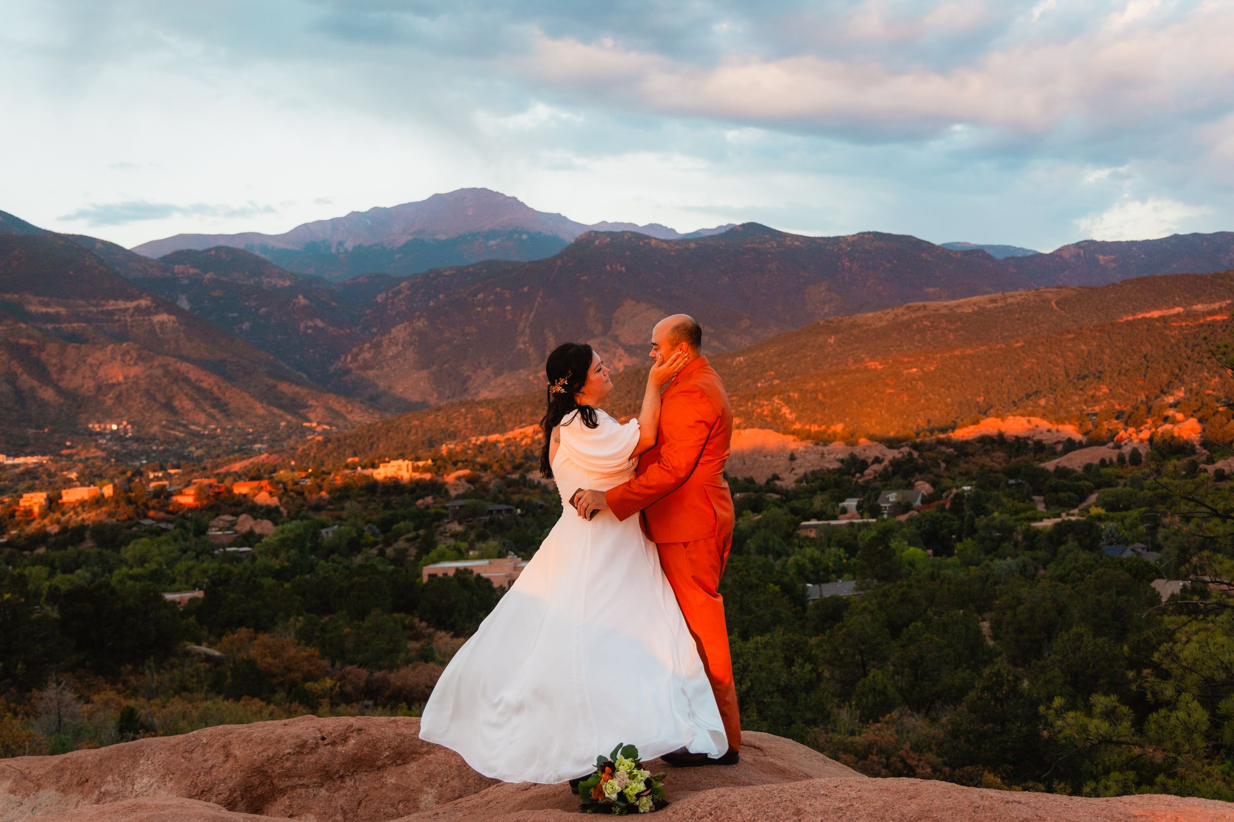 Couple in wedding attire embrace on mountain overlook at sunset.
