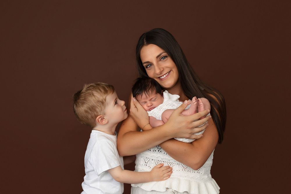A mother in a white dress holds her newborn baby while her young son kisses the infant in a studio portrait.