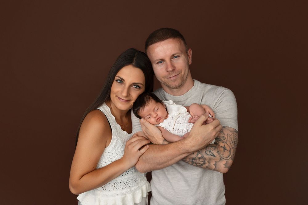 A couple smiles holding their sleeping newborn baby in a professional studio portrait against a brown background.