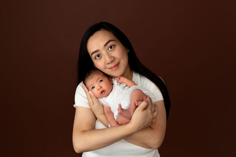 A young woman with long dark hair holds a newborn baby in a white onesie against a brown studio background.