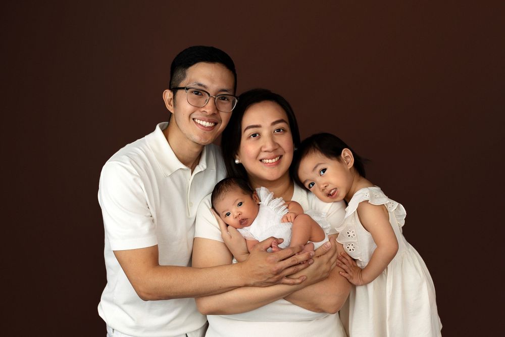 Happy Asian family portrait with parents holding newborn twins and toddler daughter, wearing white outfits on brown background.