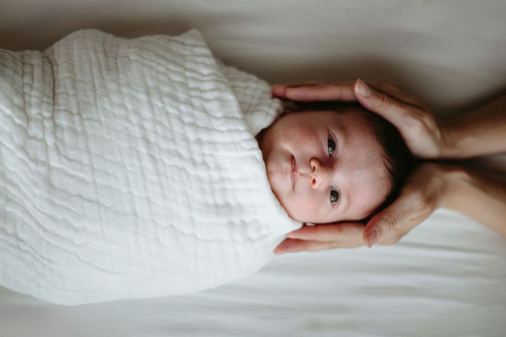 Newborn baby swaddled in white muslin cloth lying on white bed with parent's hands cradling head.